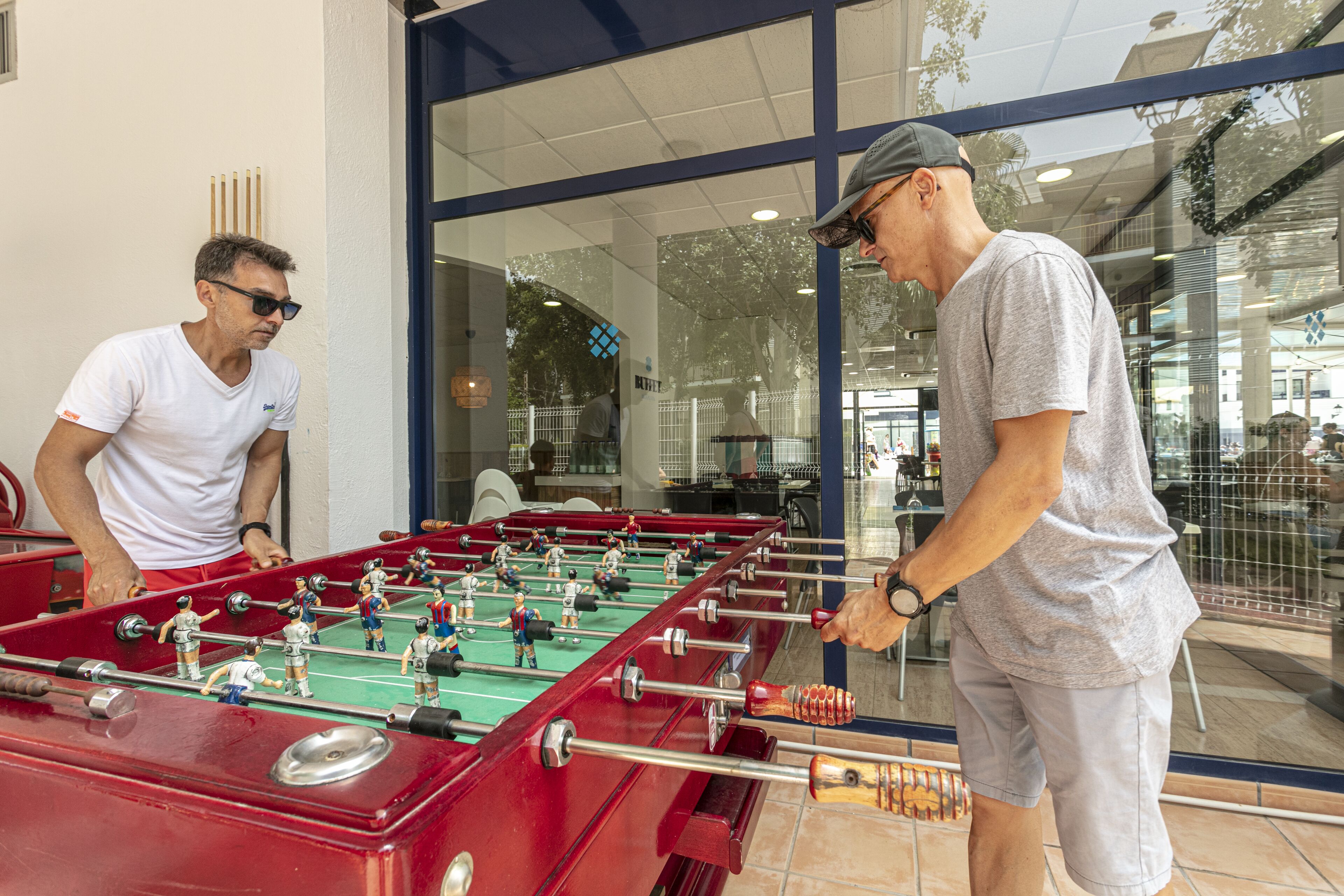 Dos hombres jugando al fútbol de mesa sobre una mesa roja en un espacio interior con ventanas amplias.