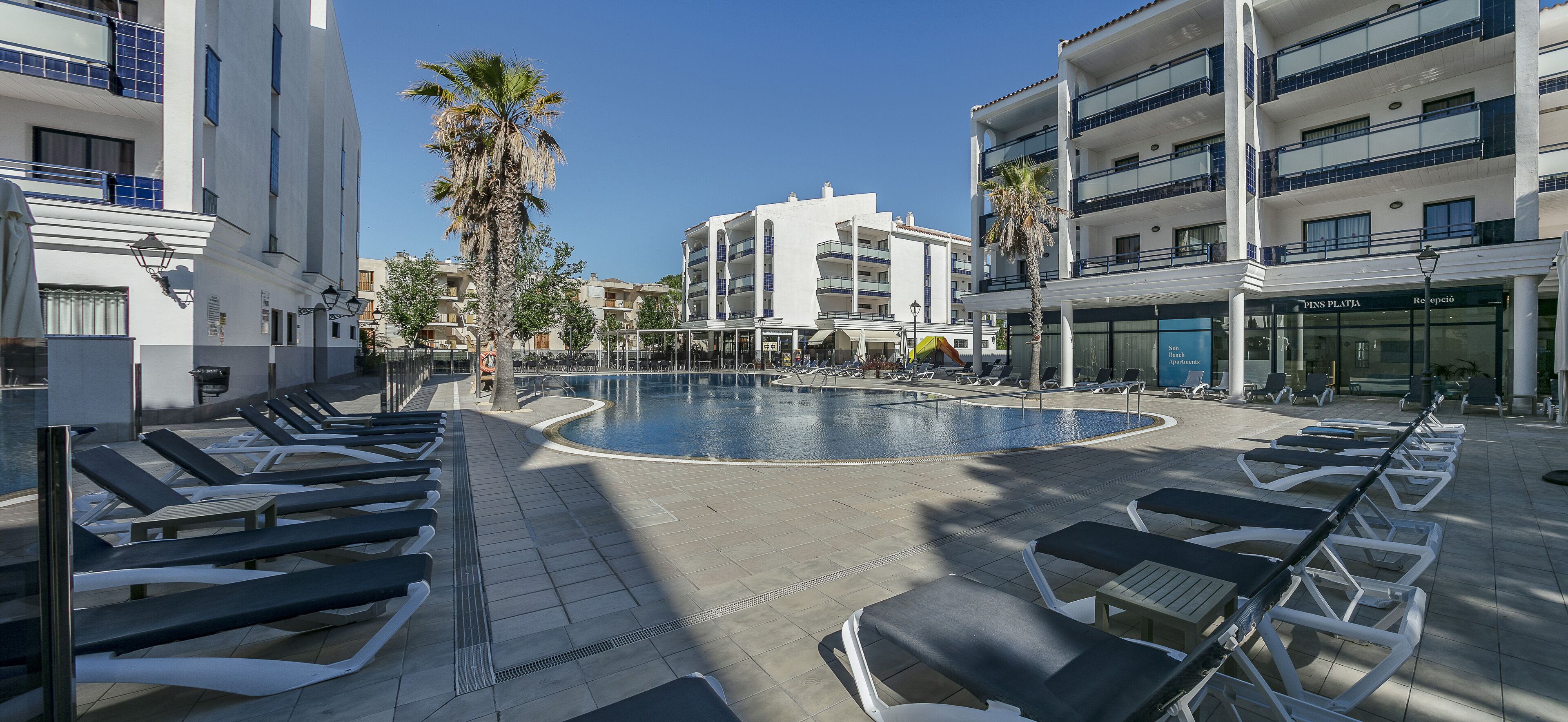 Uma área à beira da piscina com espreguiçadeiras, uma palmeira e um edifício com a placa 'PINS PLATTA'.