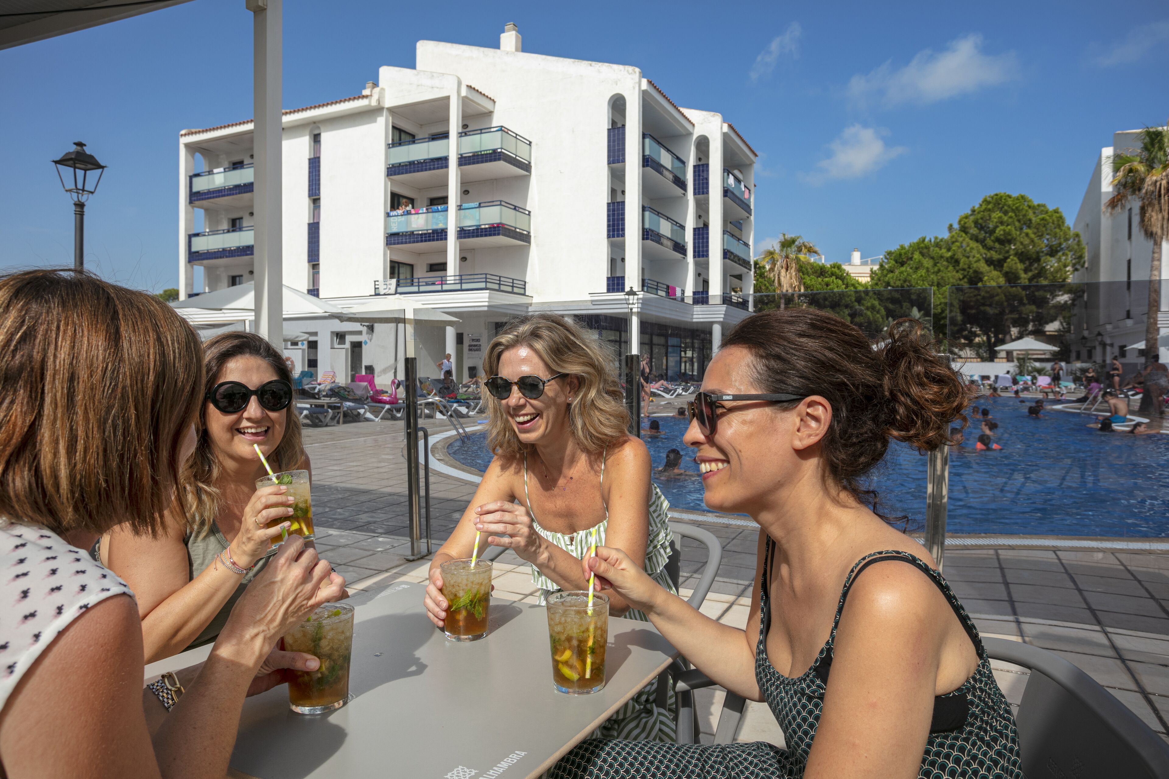 Un grup de persones assegudes a una taula al costat d'una piscina, amb un edifici al fons.