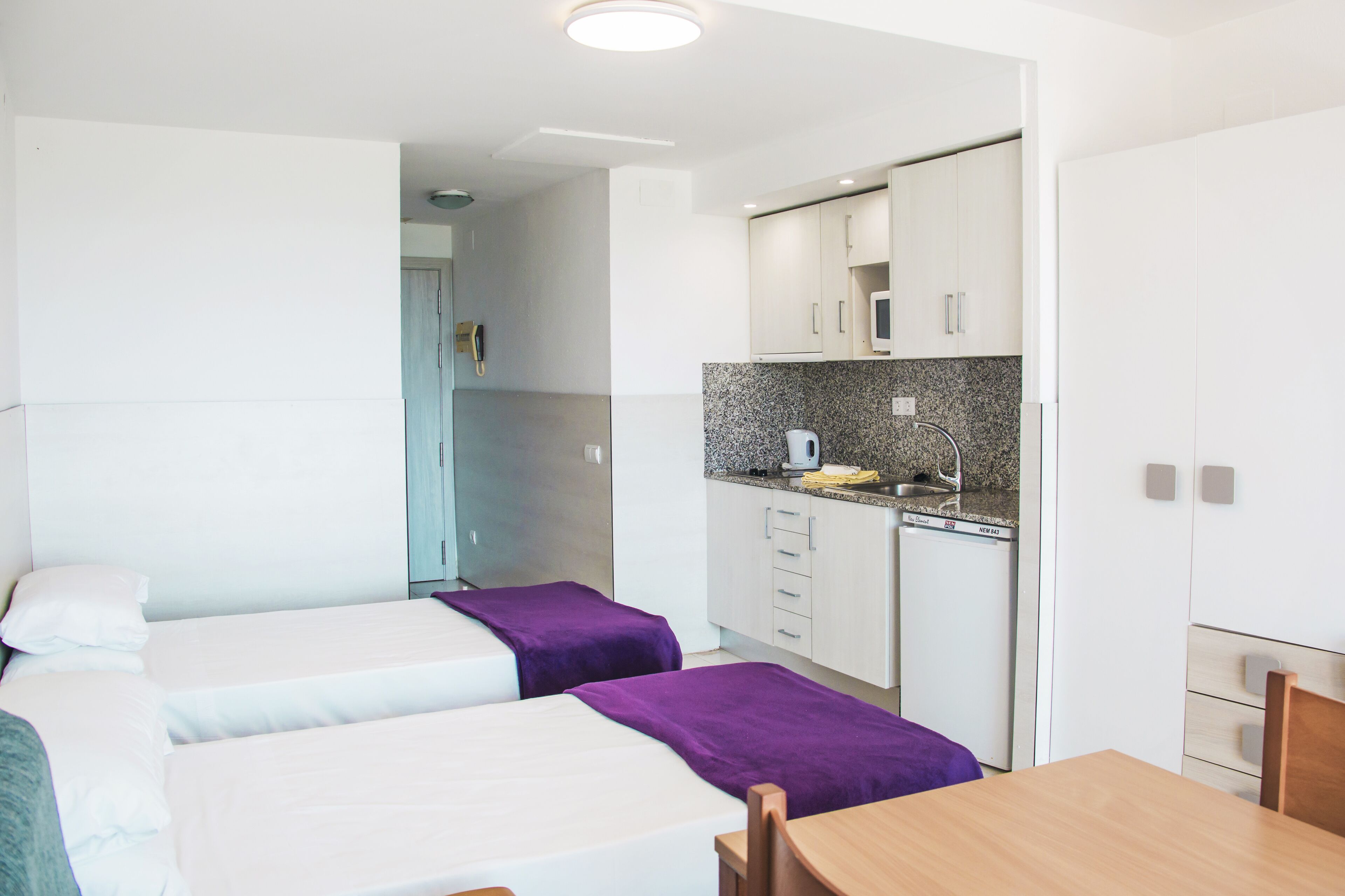 A compact hotel room with two beds, a small kitchenette, and a dining area.