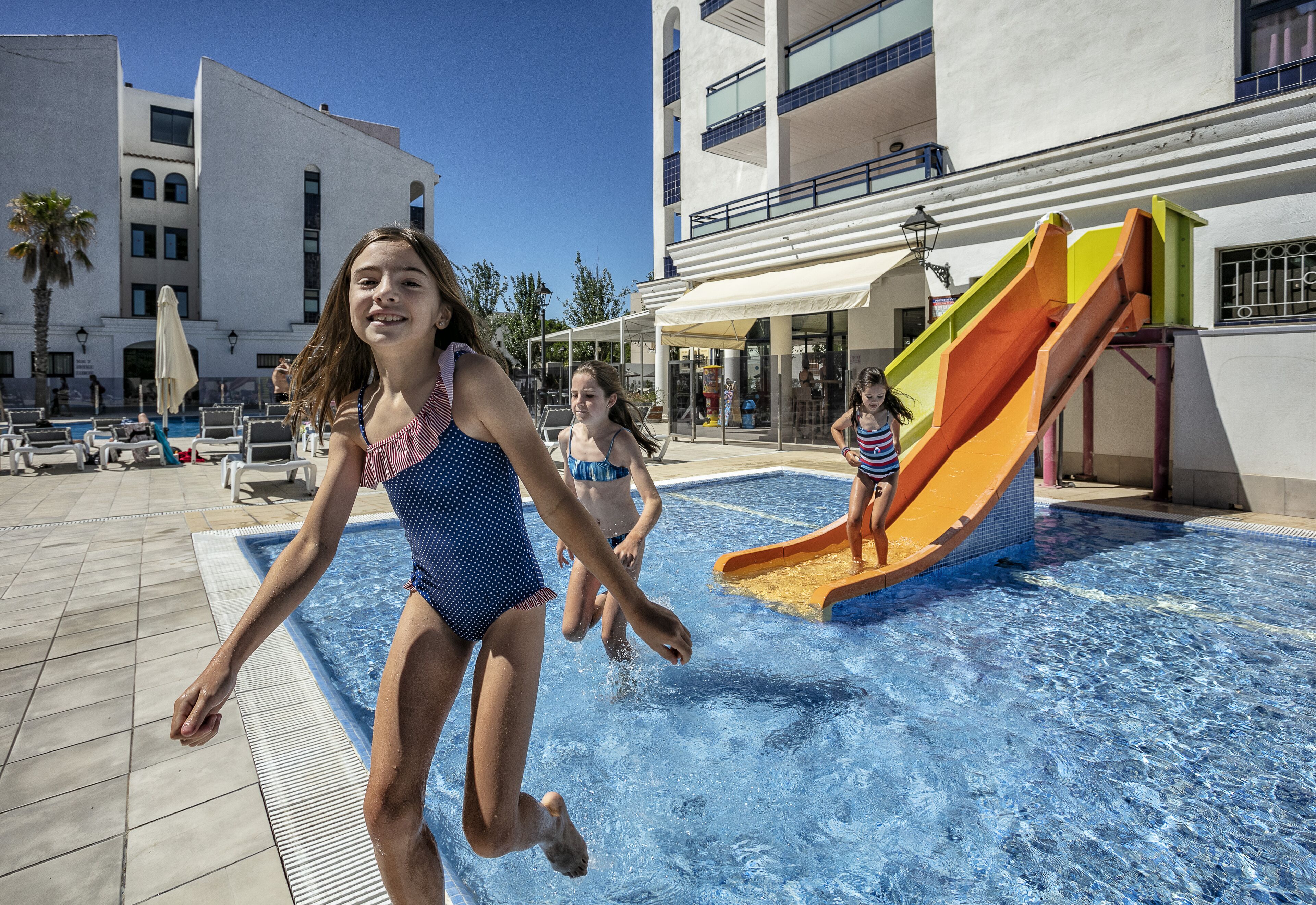 A swimming pool area with a slide, lounge chairs, and a building with balconies.