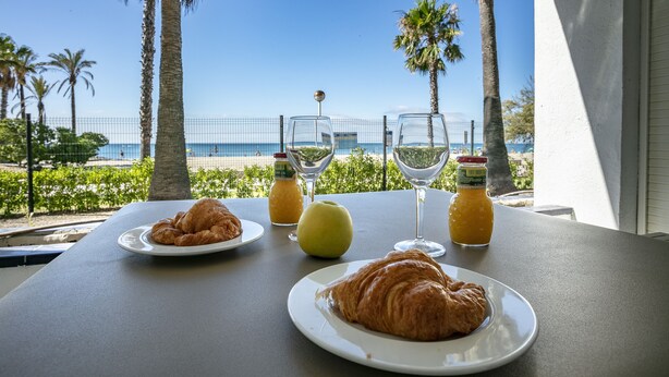 A table with croissants, a glass of juice, and a glass of white wine, overlooking a beach with palm trees and a clear blue sky.