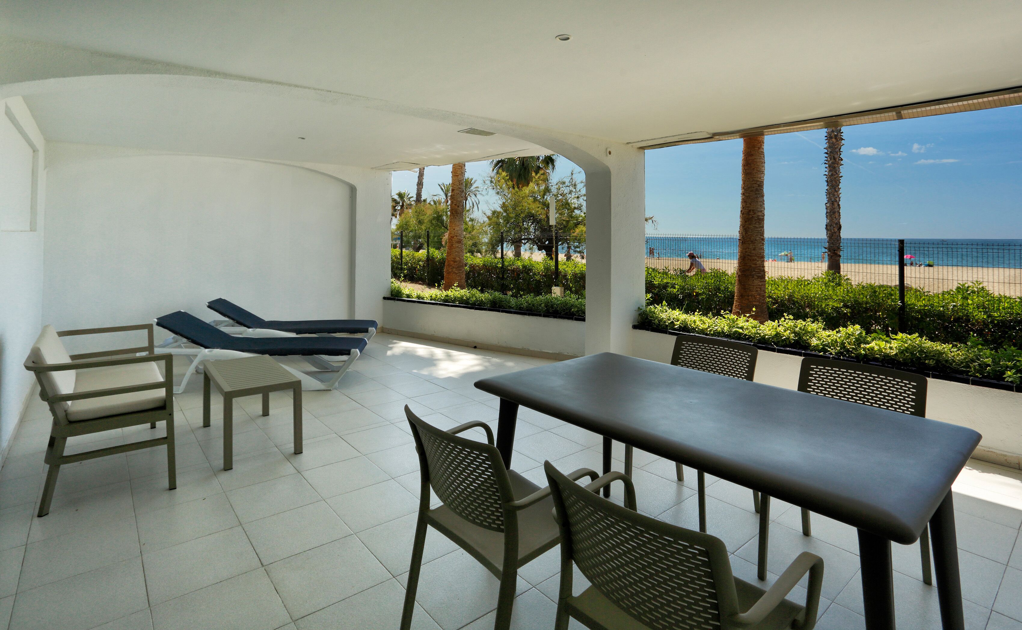 A sunlit patio with lounge chairs, a table, and chairs, overlooking a beach with palm trees and a fence.
