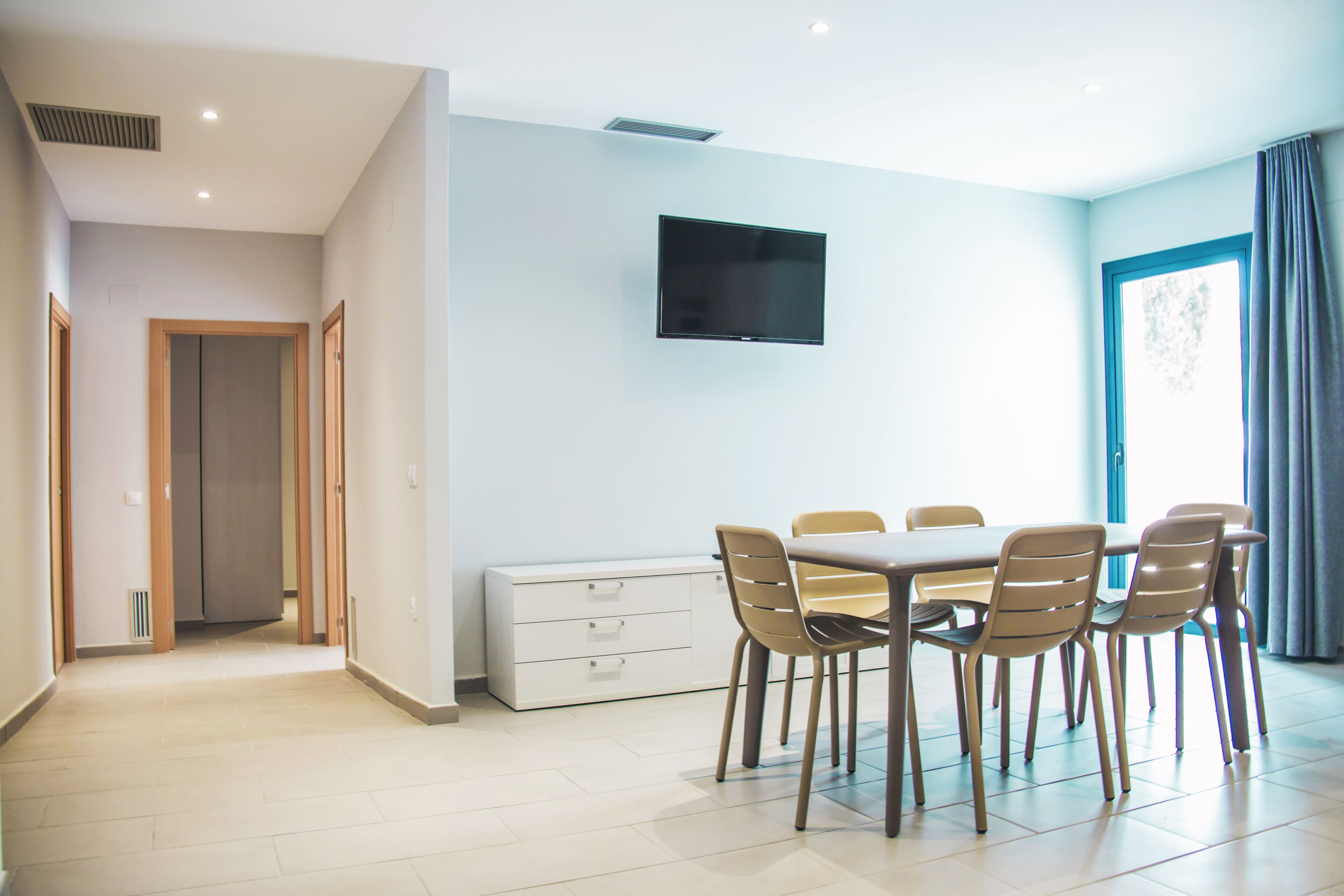Un comedor moderno con una mesa y sillas, un televisor de pantalla plana en la pared y vistas al exterior a través de una ventana.