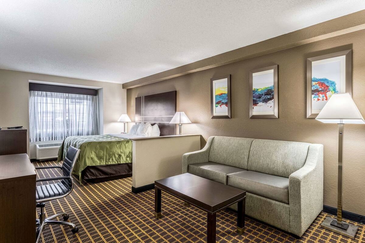 A hotel room with a bed, a sofa, a desk, a lamp, and artwork on the wall.
