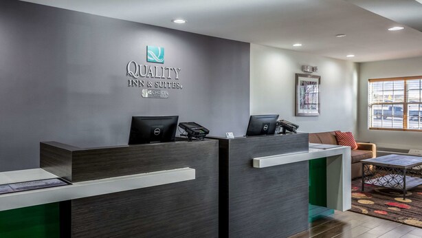 Een receptiebalie van een hotel met een logo, twee computerschermen, een telefoon en een zithoek met een bank en een raam.