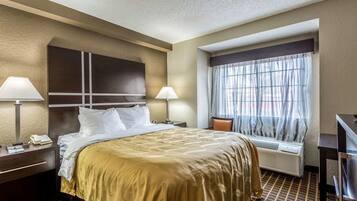 A hotel room with a large bed, two bedside lamps, a window with sheer curtains, a built-in air conditioning unit, and a dark wooden nightstand with a phone and papers.