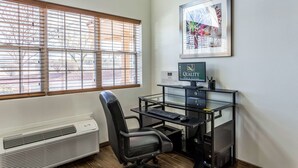 Een moderne kantoorruimte met een bureau, stoel en computer. Er is een raam met zonwering, een lijst aan de muur en een bord met daarop ‘quality inn and suites’.