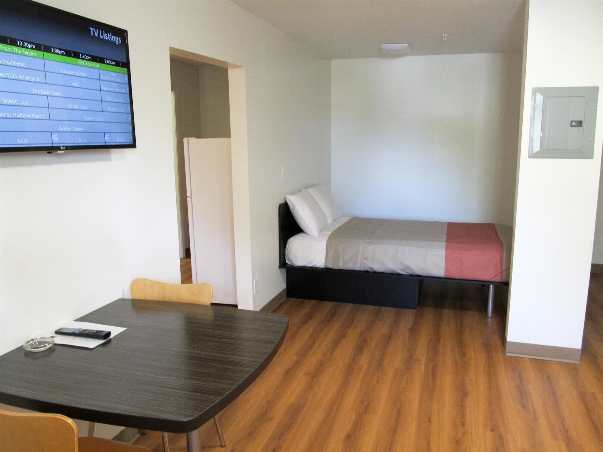 A small, modern room with a wooden floor, a bed with a red and white blanket, a desk with a chair, and a TV displaying TV listings.