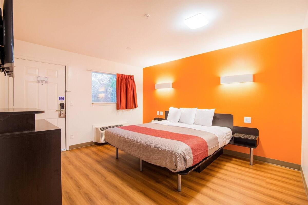 A hotel room with a bed, a desk, a window with curtains, and an orange accent wall.