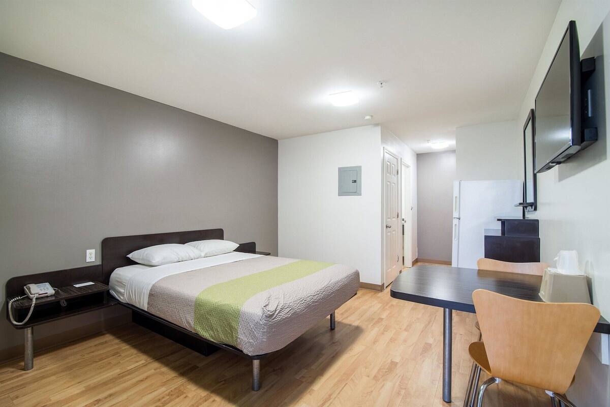 A compact hotel room with a bed, bedside table, chair, desk, and a TV mounted on the wall.