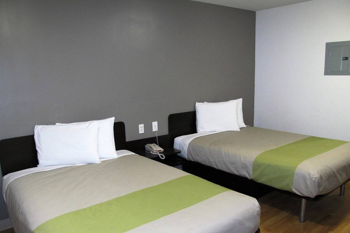 A hotel room with two single beds, each with a white pillow and a green accent on the bedspread. There is a bedside table with a telephone and a power outlet on the wall.