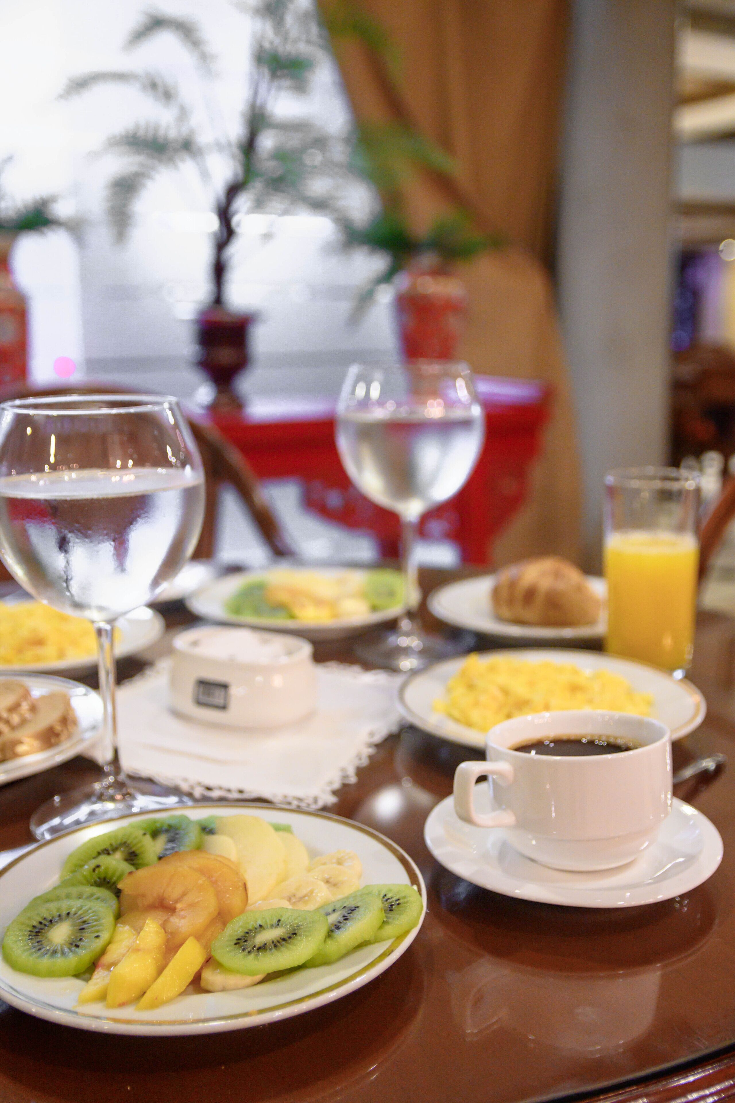 A breakfast table with plates of fruit, a glass of water, a glass of wine, a cup of coffee, and a glass of orange juice.