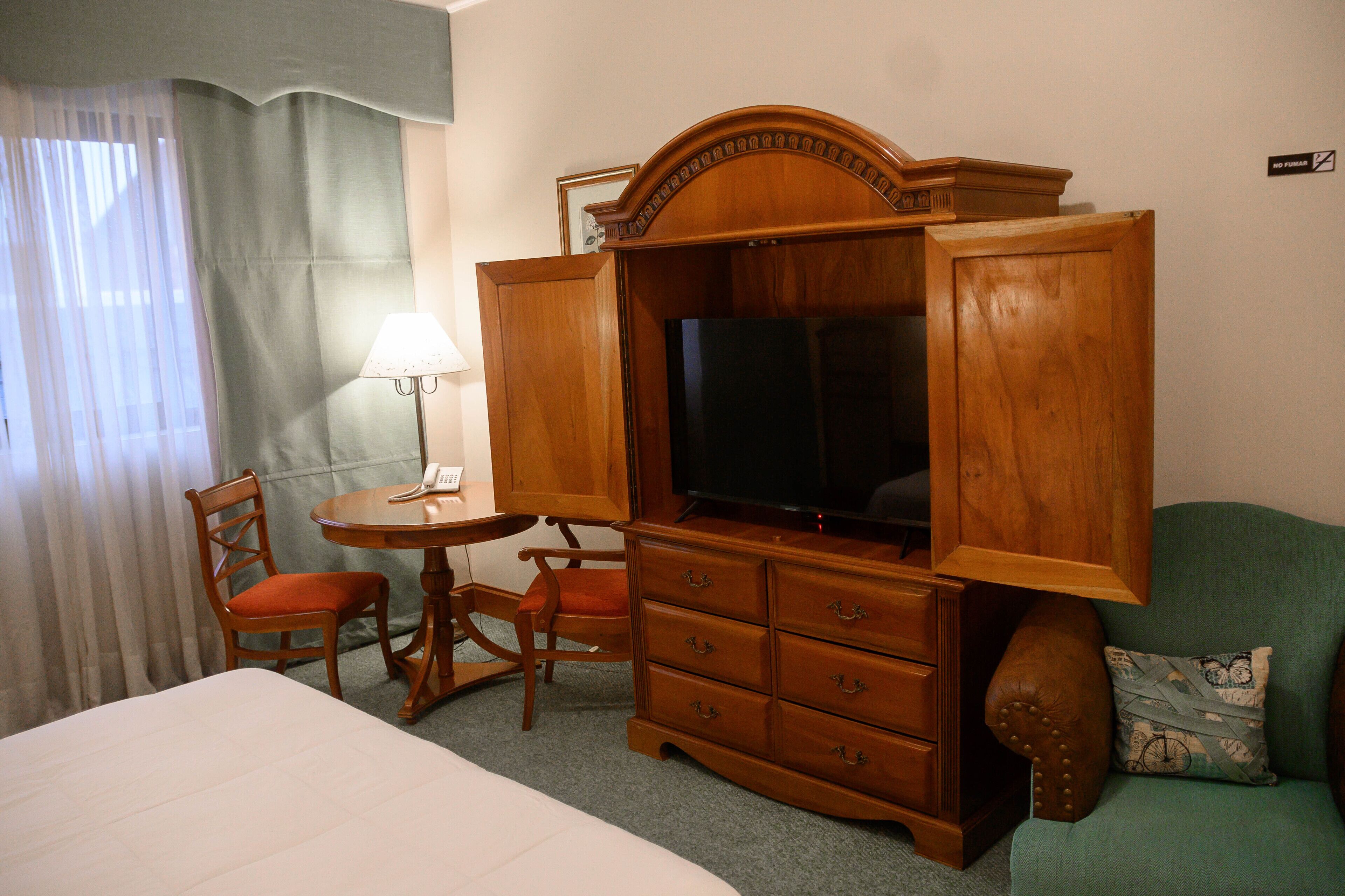 A hotel room with a bed, a wooden TV stand with a television, a desk with a lamp, two chairs, and a green sofa.