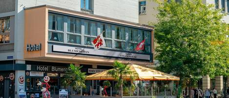 Un hotel con fachada moderna, un letrero llamativo del 'Hotel Blumenstein' y 'René's Steakhouse', y un área de estar al aire libre cubierta con palmeras.