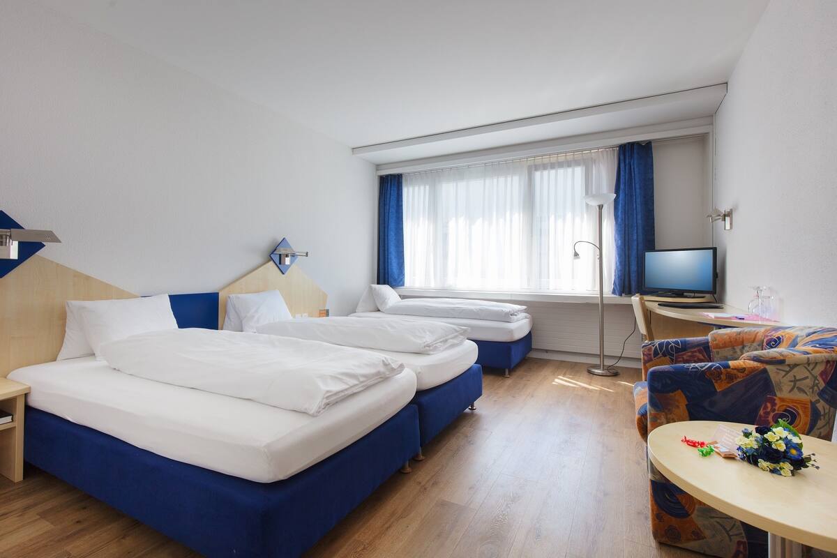 A hotel room with two beds, a desk, a television, and a small table with flowers.