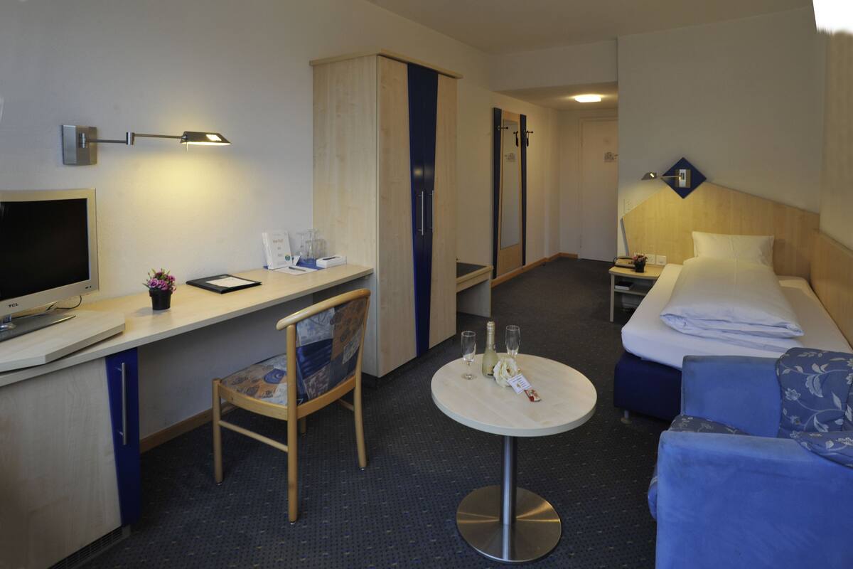 A hotel room with a desk, chair, bed, bedside table, and a small round table with a bottle and glasses.