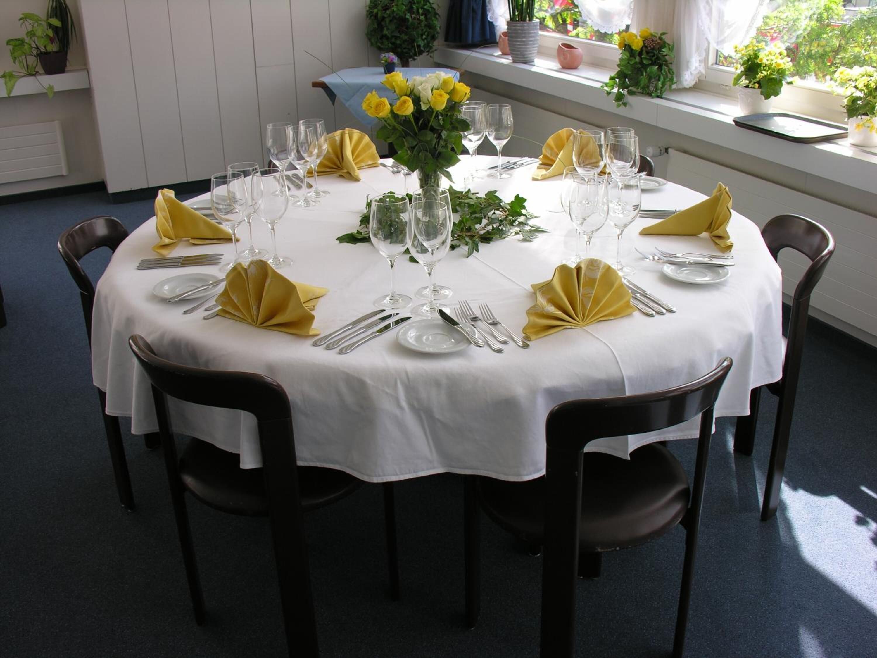 A round dining table set for six with white tablecloths, yellow napkins, and a vase of yellow flowers.