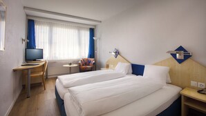 Hotellihuone, jossa on kaksi sänkyä, työpöytä, jossa on tietokone, tuoli, televisio ja ikkuna, jossa on verhot.