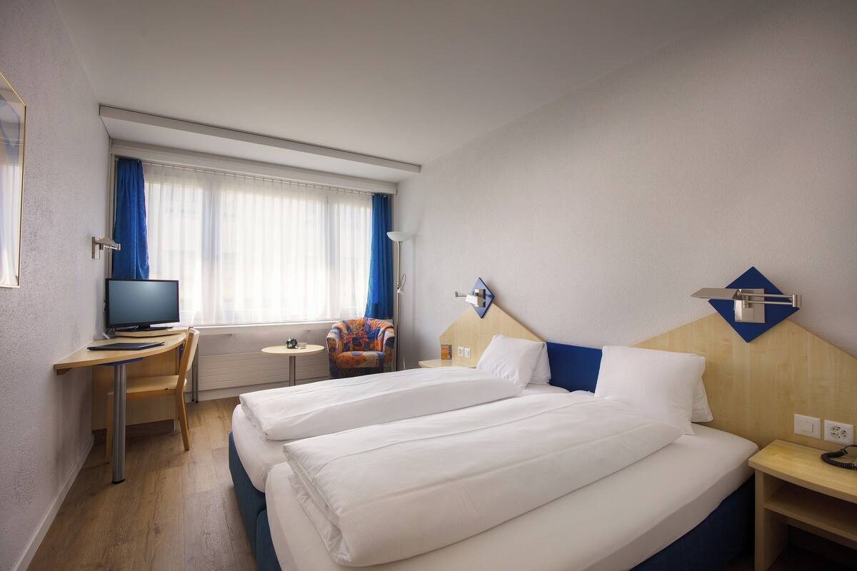 A hotel room with two beds, a desk with a computer, a chair, a TV, and a window with curtains.
