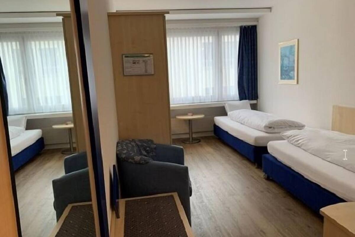 A hotel room with two beds, a small table, a chair, and a window with curtains.