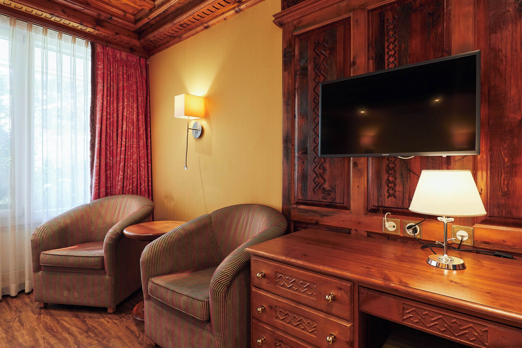 A hotel room with a wooden desk, two striped armchairs, a flat-screen TV, and a lamp.