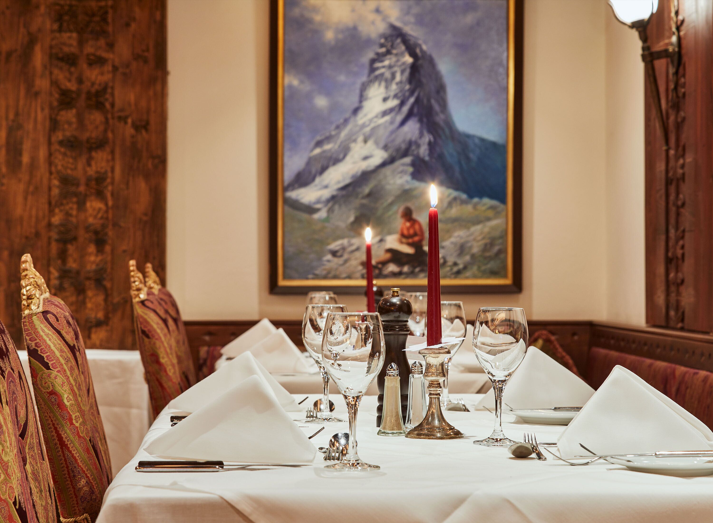A dining table set with white tablecloth, red candles, and glassware. A painting of a mountain is on the wall behind.