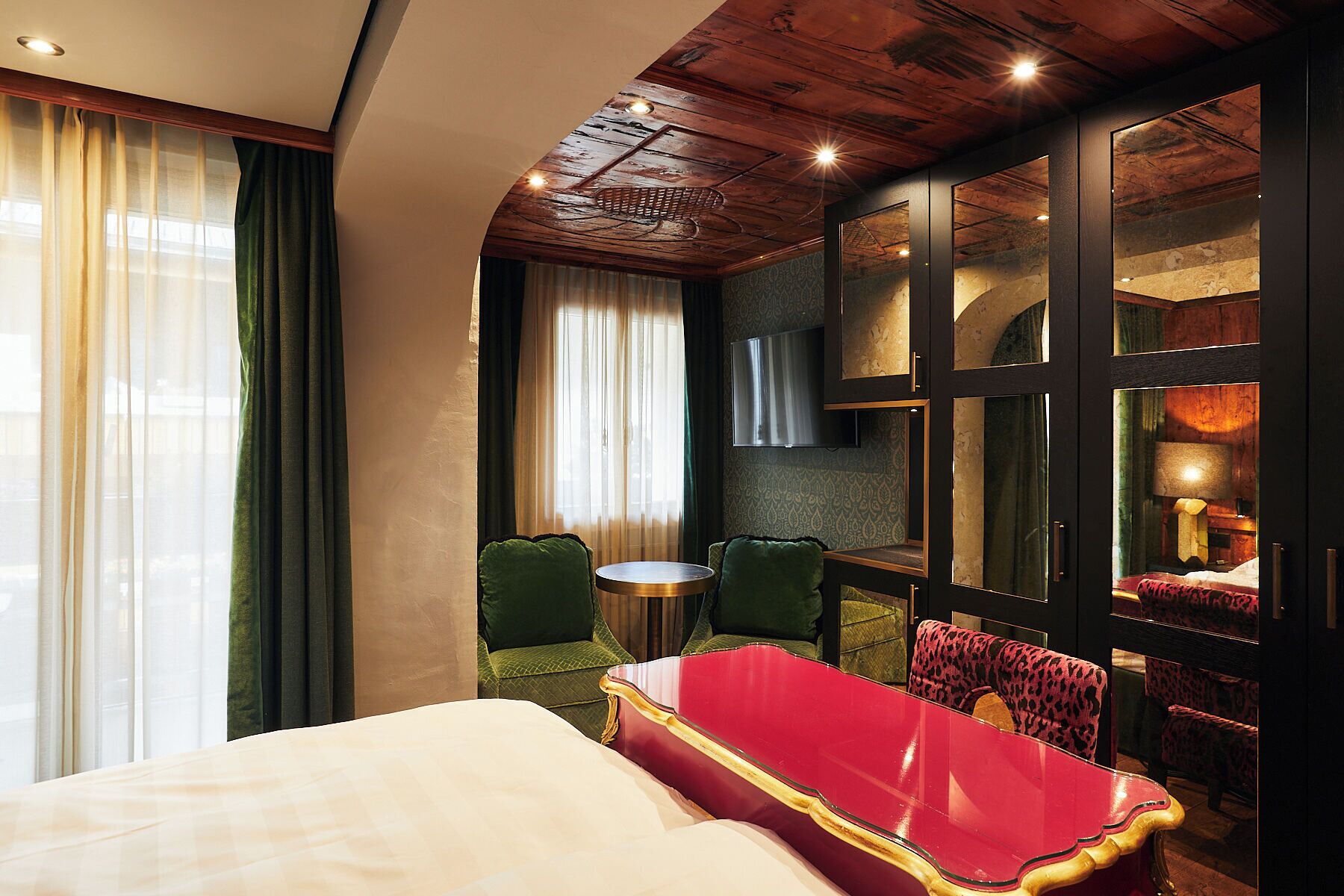Ein Hotelzimmer mit einem Bett, einem roten Sessel, einem grünen Sessel, einem runden Tisch und einem an der Wand montierten Fernseher.