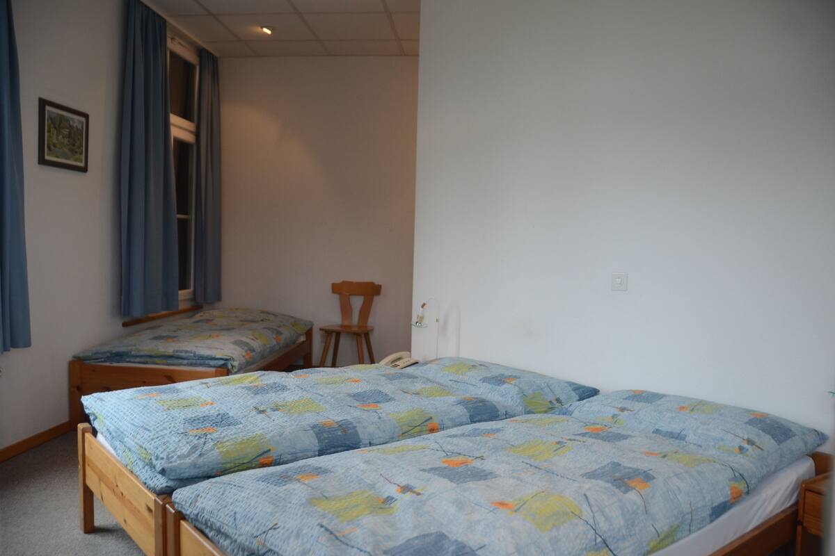 A room with two wooden beds, a chair, a window with blue curtains, and a wall-mounted telephone.