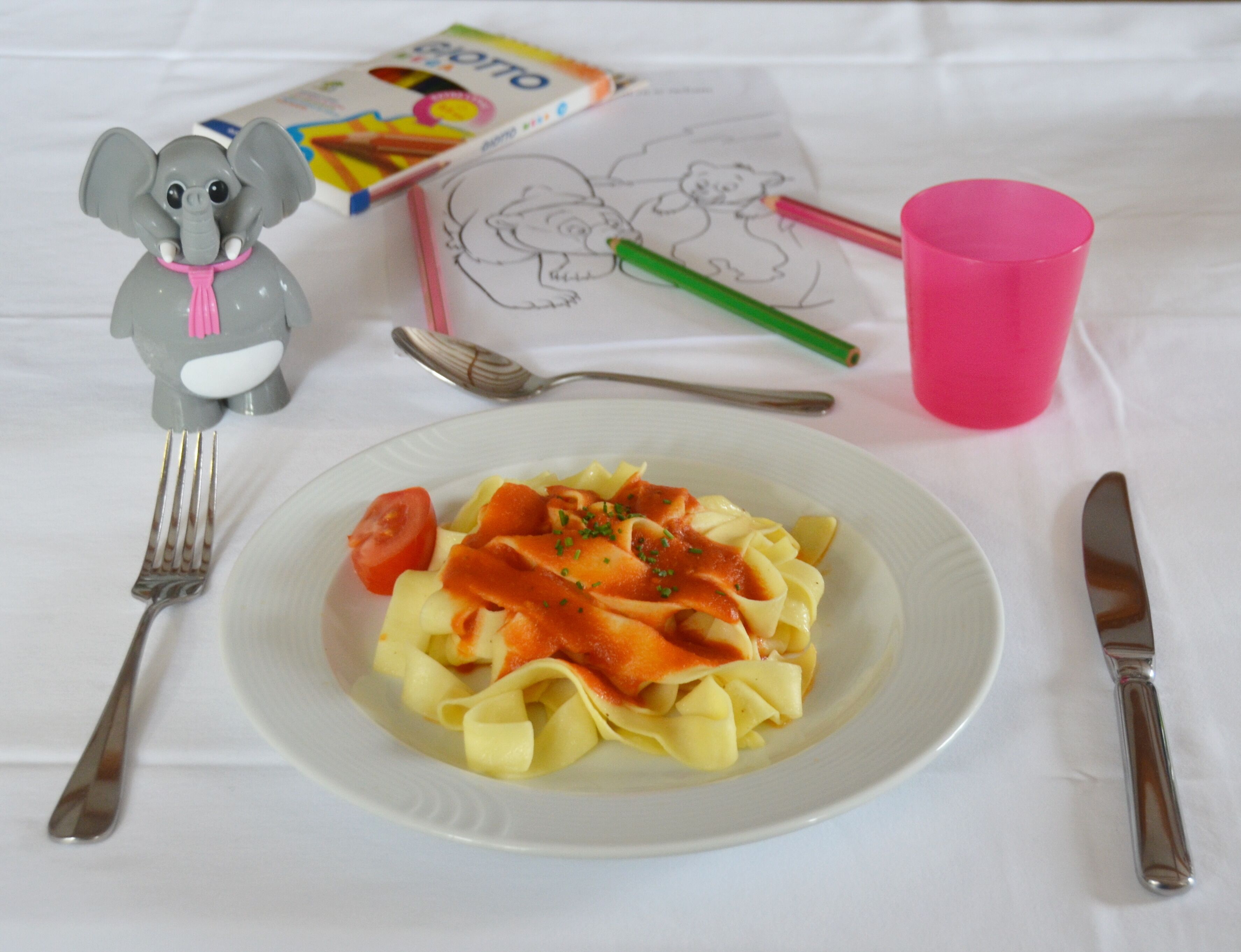 Eine Pasta-Kombination mit Tomatensauce und einer Kirschtomate, eine Gabel, ein Messer, ein Löffel, eine rosa Tasse, ein Bleistift und ein Malbuch mit einer Bär-Zeichnung.