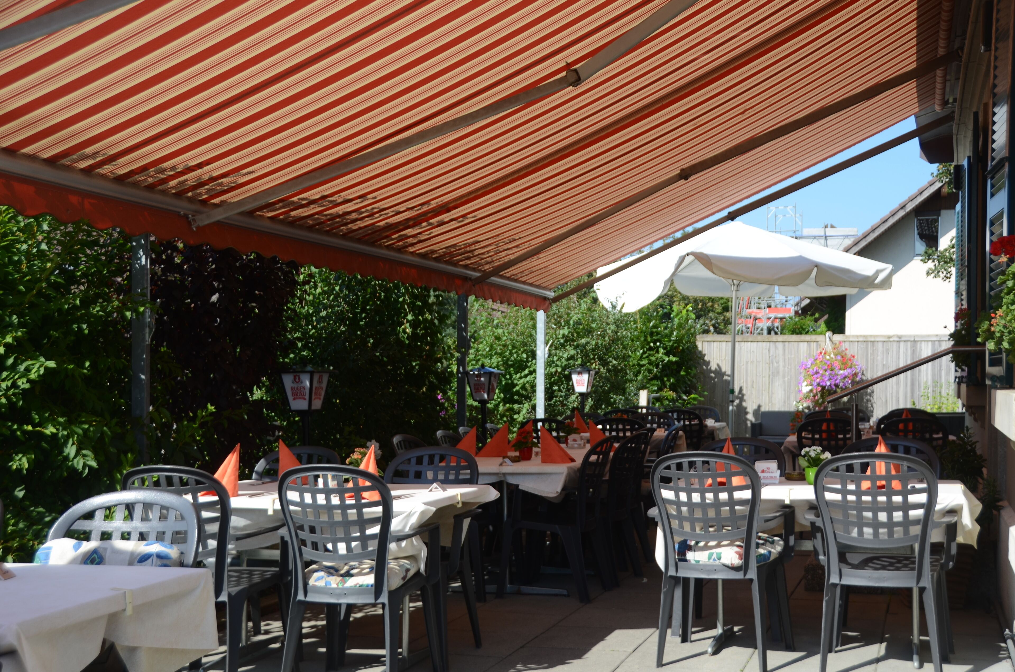Une terrasse extérieure avec des tables et des chaises, protégée par un auvent à rayures et entourée de verdure.