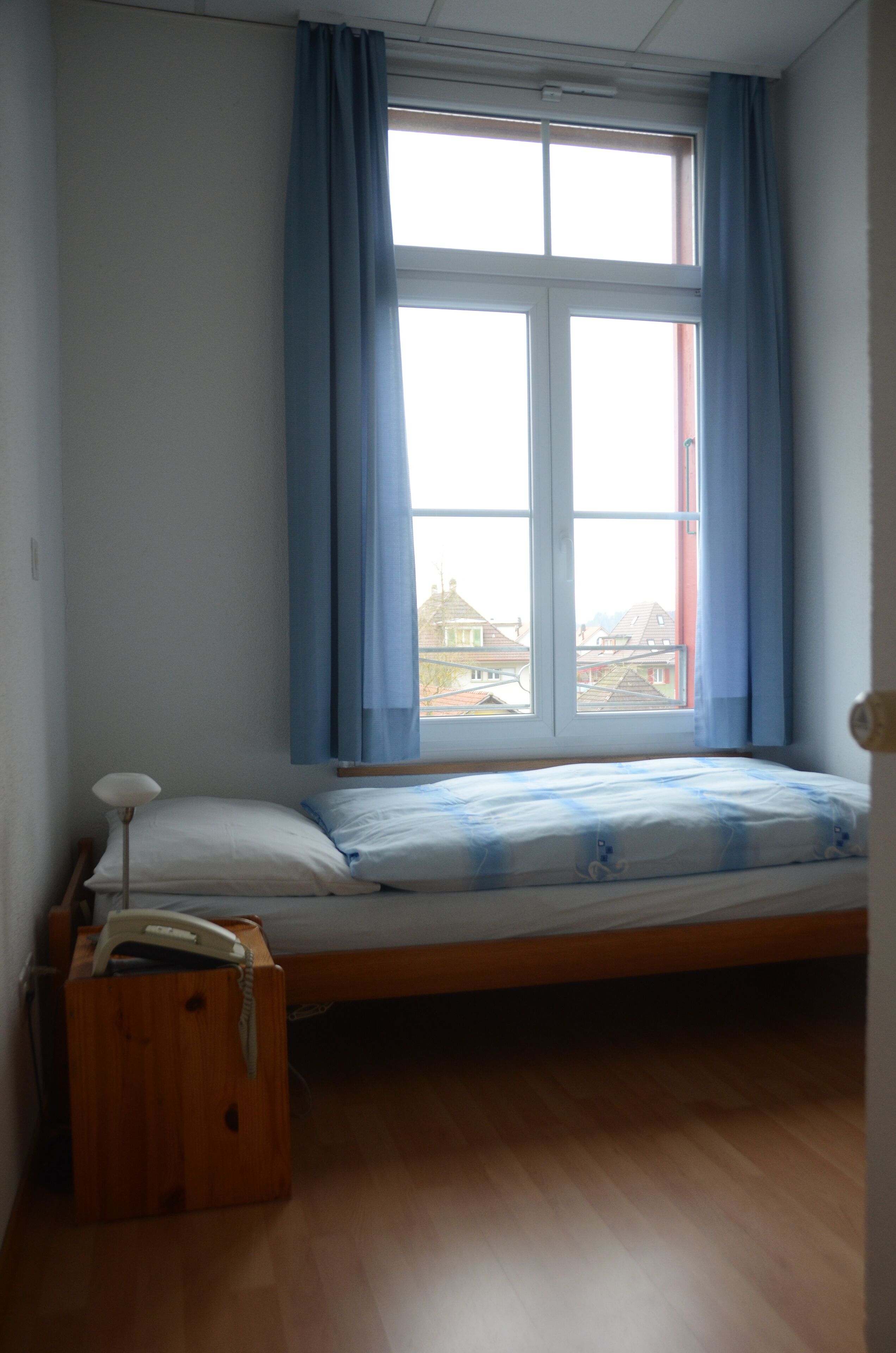 A single bed with a wooden frame, a bedside table with a lamp, and a window with blue curtains.
