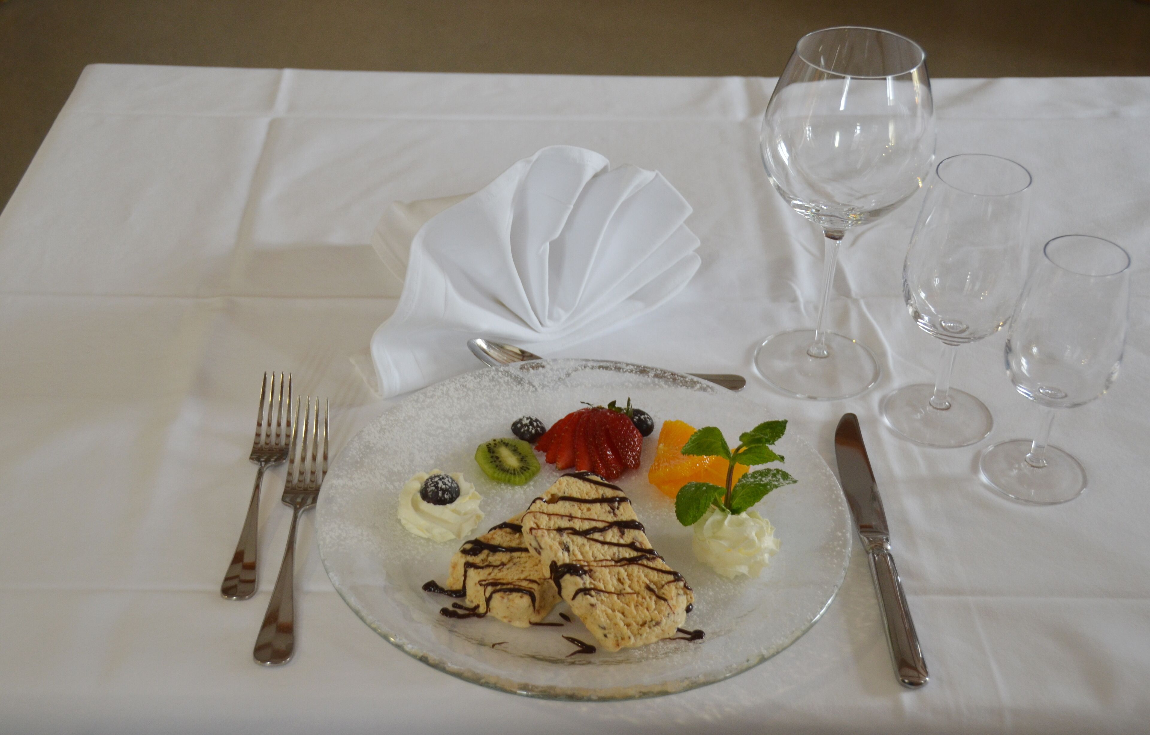 Ein Dessertteller mit verschiedenen Früchten und einem Gebäck auf einer weißen Tischdecke, begleitet von Besteck und Weingläsern.