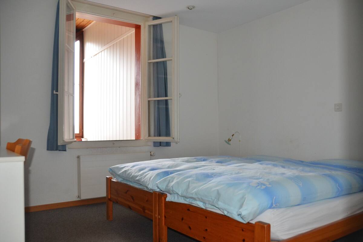 A bedroom with a single bed, a window with curtains, a radiator, and a small desk with a chair.