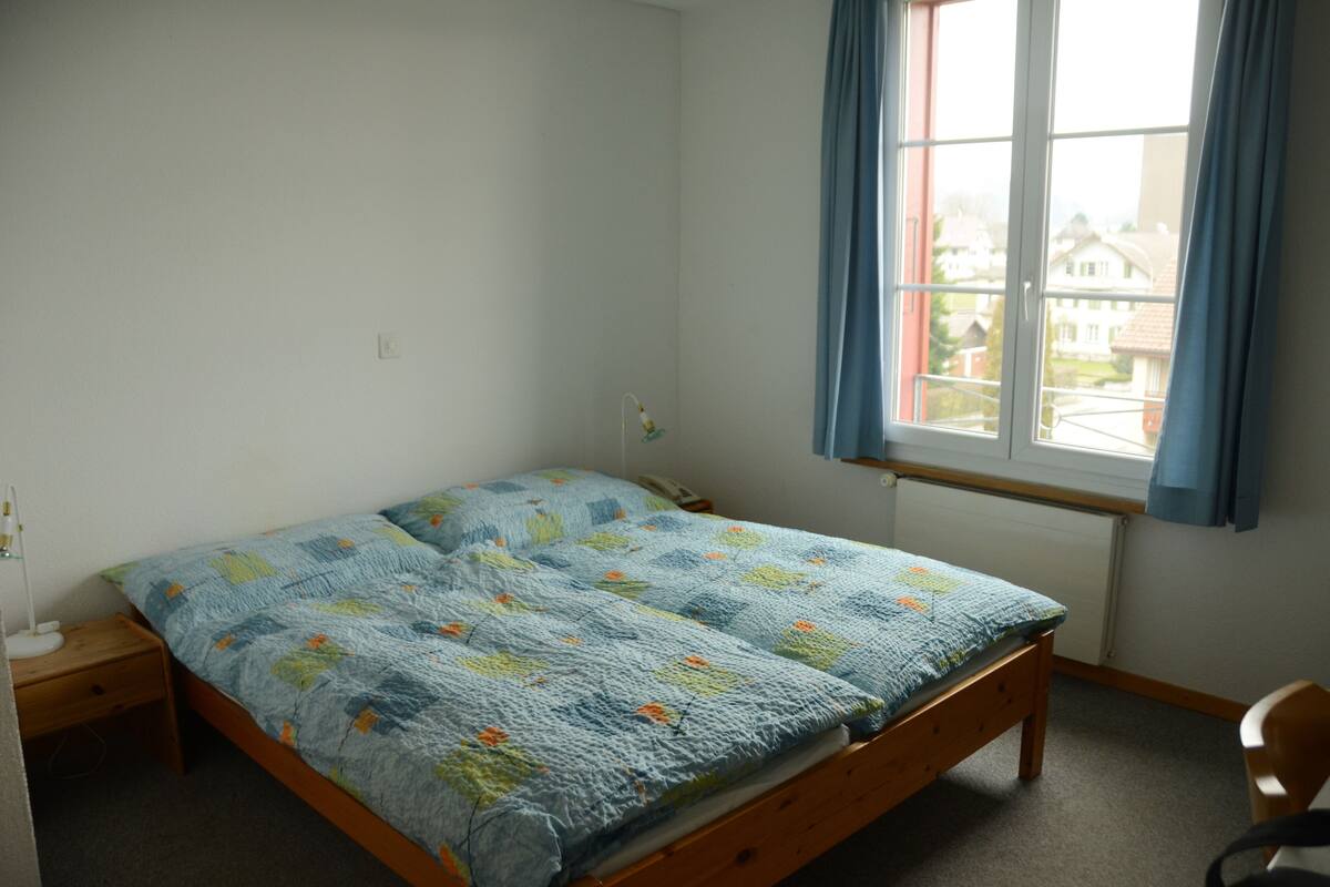 A single bed with a blue floral bedspread in a room with white walls, a window with blue curtains, and a wooden nightstand with a lamp.