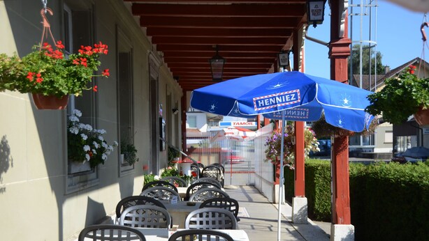 An outdoor cafe with tables and chairs, a blue umbrella, hanging flower baskets, and a building with a sign. - Hotel Bahnhof (Schupfen)