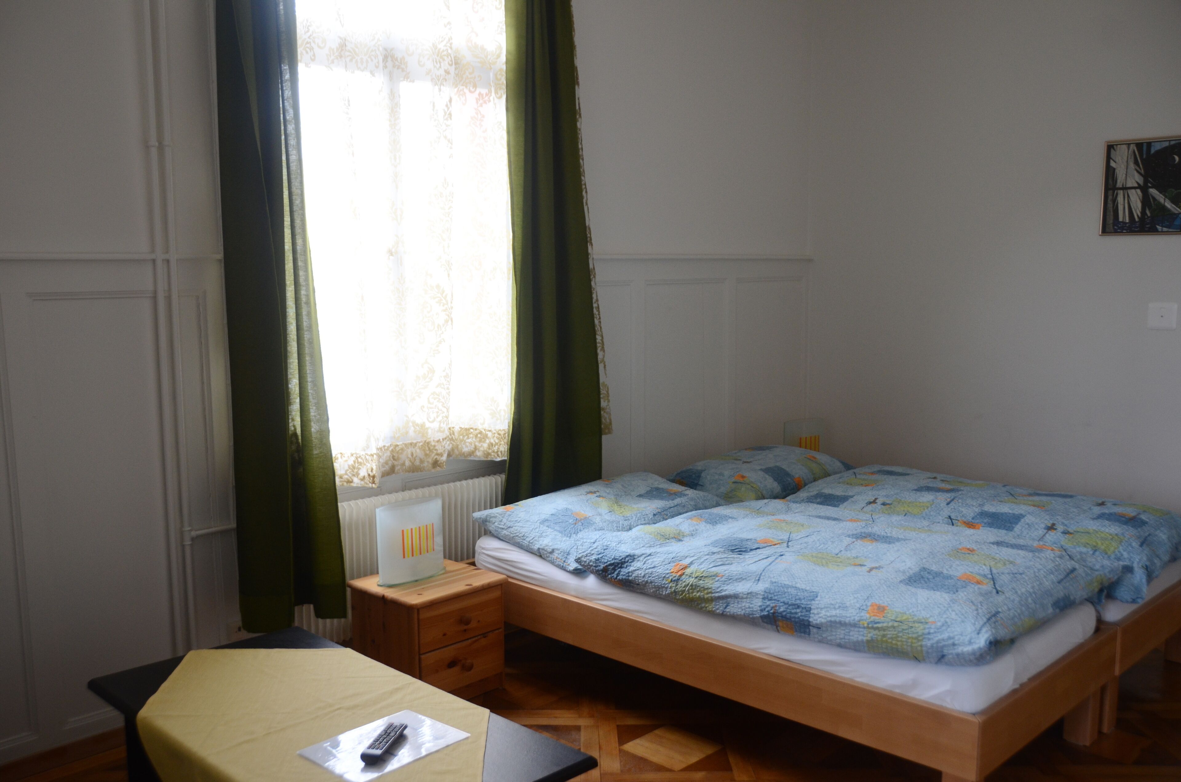 A single bed with a blue quilt, a bedside table with a lamp, a chair, and a window with green curtains.