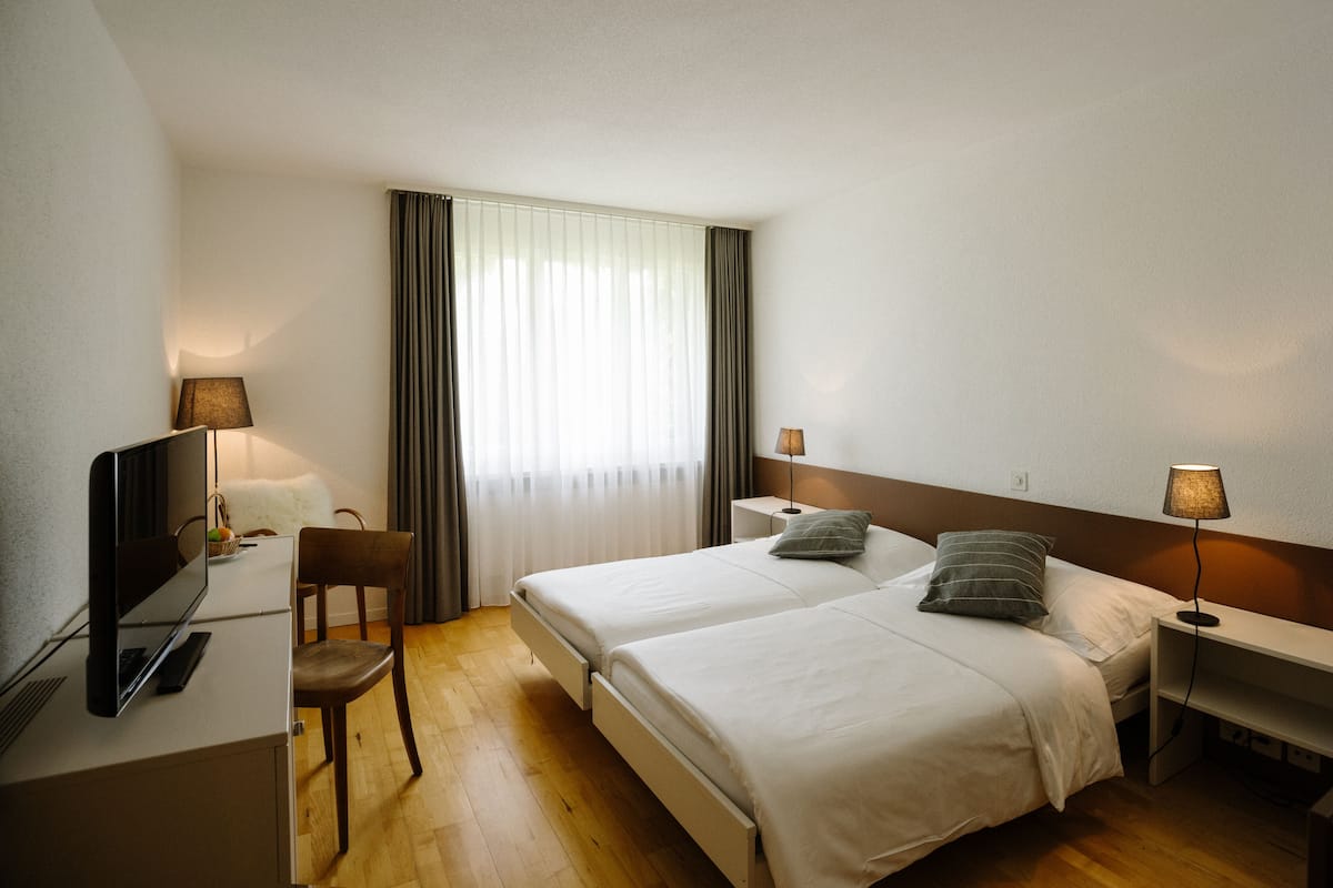 A hotel room with two beds, a desk with a TV, a chair, and a window with curtains.