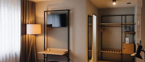 A hotel room with a bed, a desk, a mirror, a TV, a wardrobe, and a shelf with toiletries.