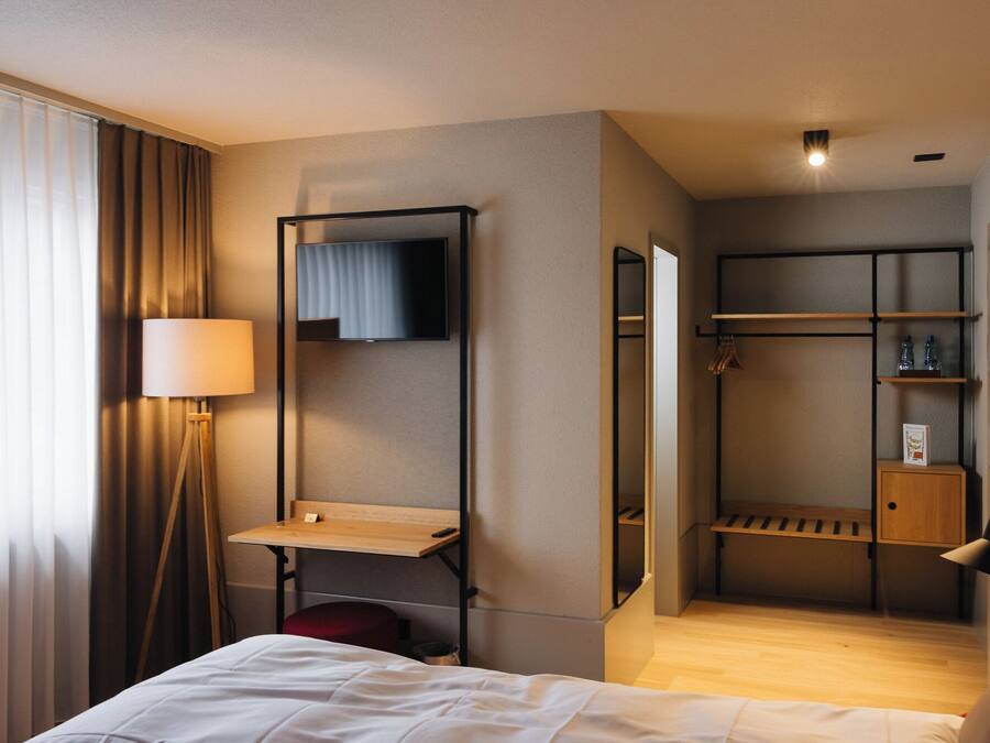 A hotel room with a bed, a desk, a mirror, a TV, a wardrobe, and a shelf with toiletries.