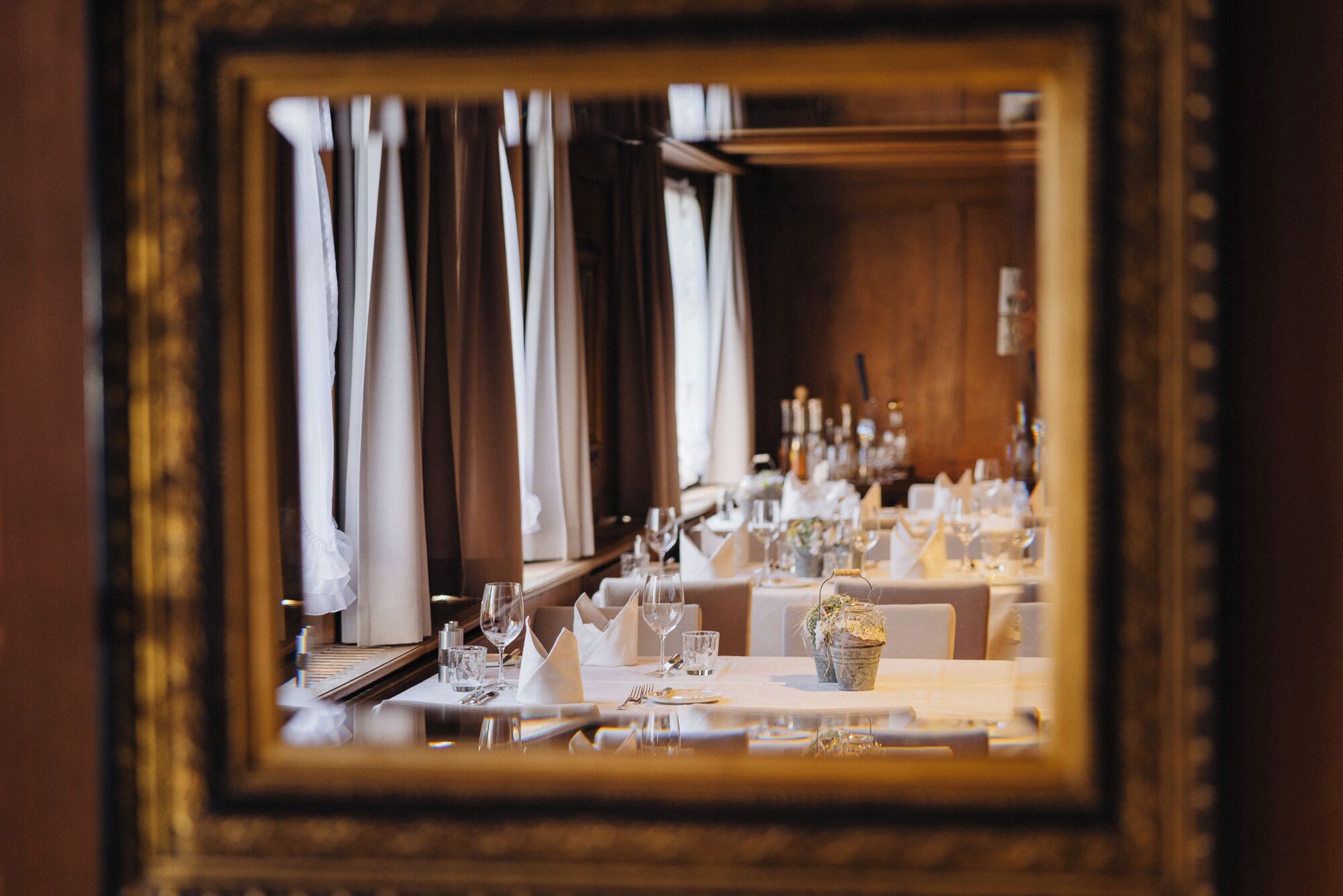 Un comedor con mesas dispuestas para una comida, un espejo que refleja la habitación y cortinas en las ventanas.