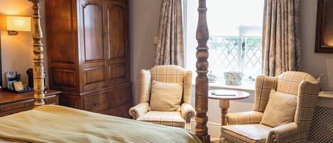 Un dormitorio con una cama con dosel, dos sillones a cuadros, un armario de madera y una ventana con cortinas.