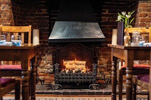 A fireplace with a log fire, surrounded by wooden furniture and a brick wall. - Red Lion Hotel, Hillingdon (Uxbridge)
