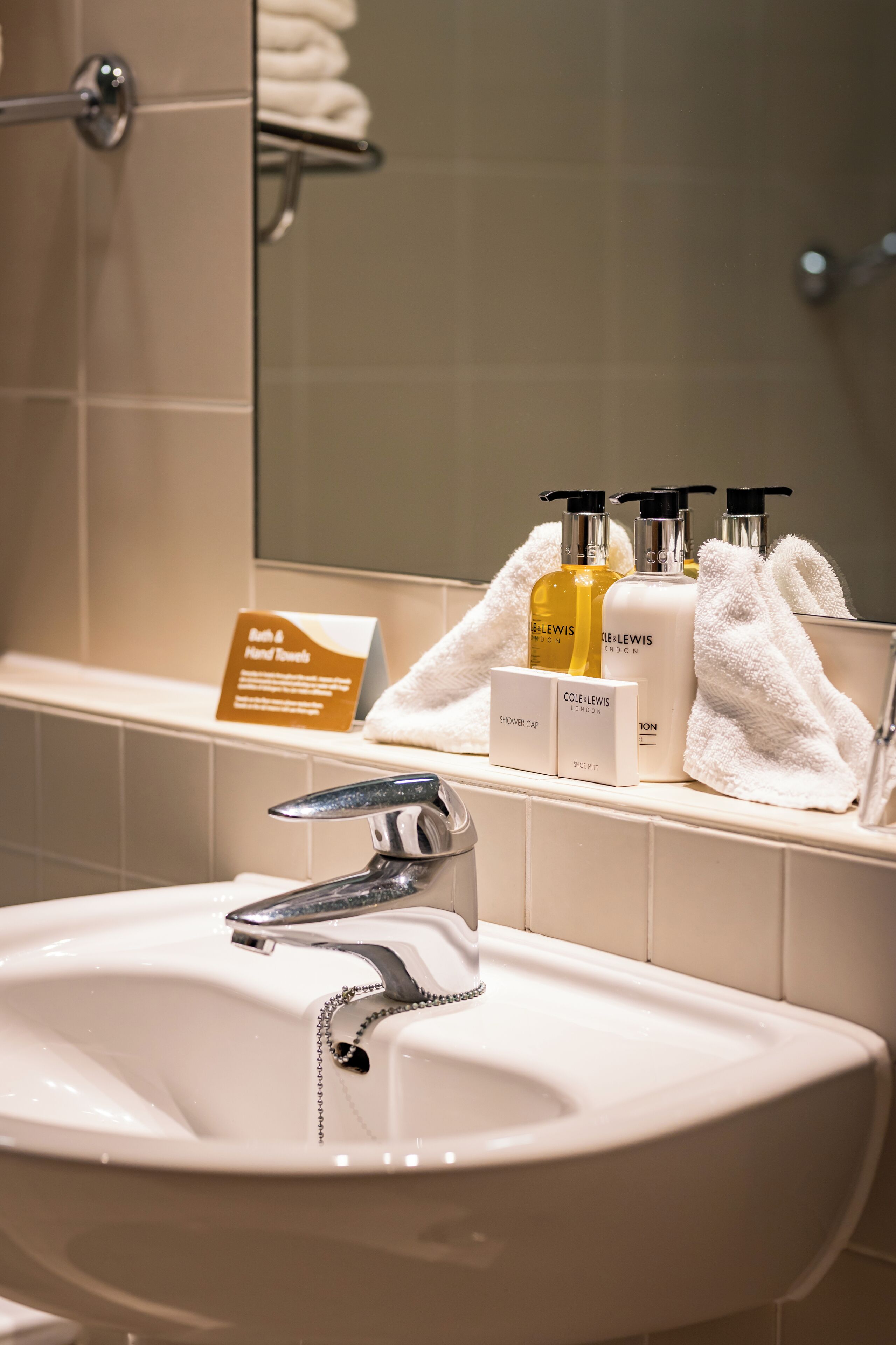 A bathroom with a white sink, chrome faucet, and a mirror above it. There are toiletries on the shelf, including bottles labeled 'Cole Lewis London' and 'Bath & Hand Soap'.