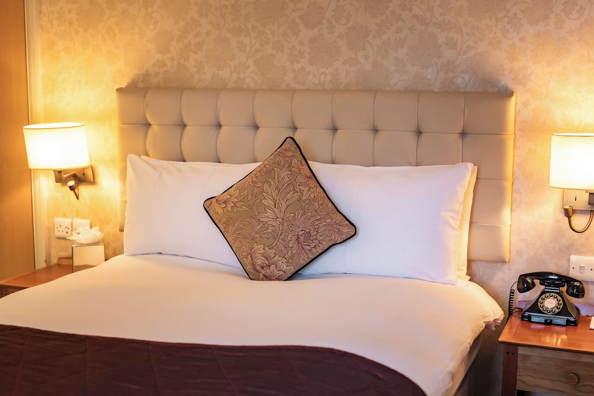 A hotel room with a tufted headboard, a white bedspread, a decorative pillow, a bedside telephone, and wall-mounted lamps.