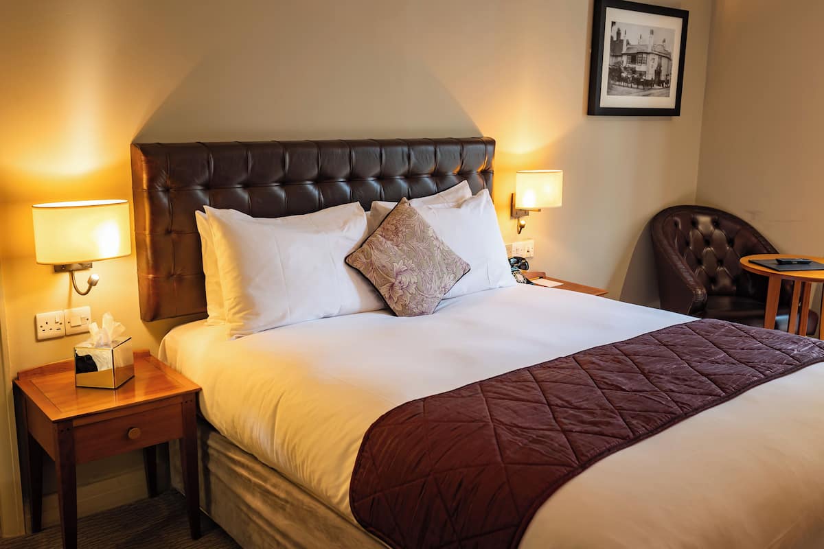 A neatly made bed with a dark headboard, white linens, and a brown quilt. There are two bedside lamps, a nightstand with a tissue box, and a framed picture on the wall.