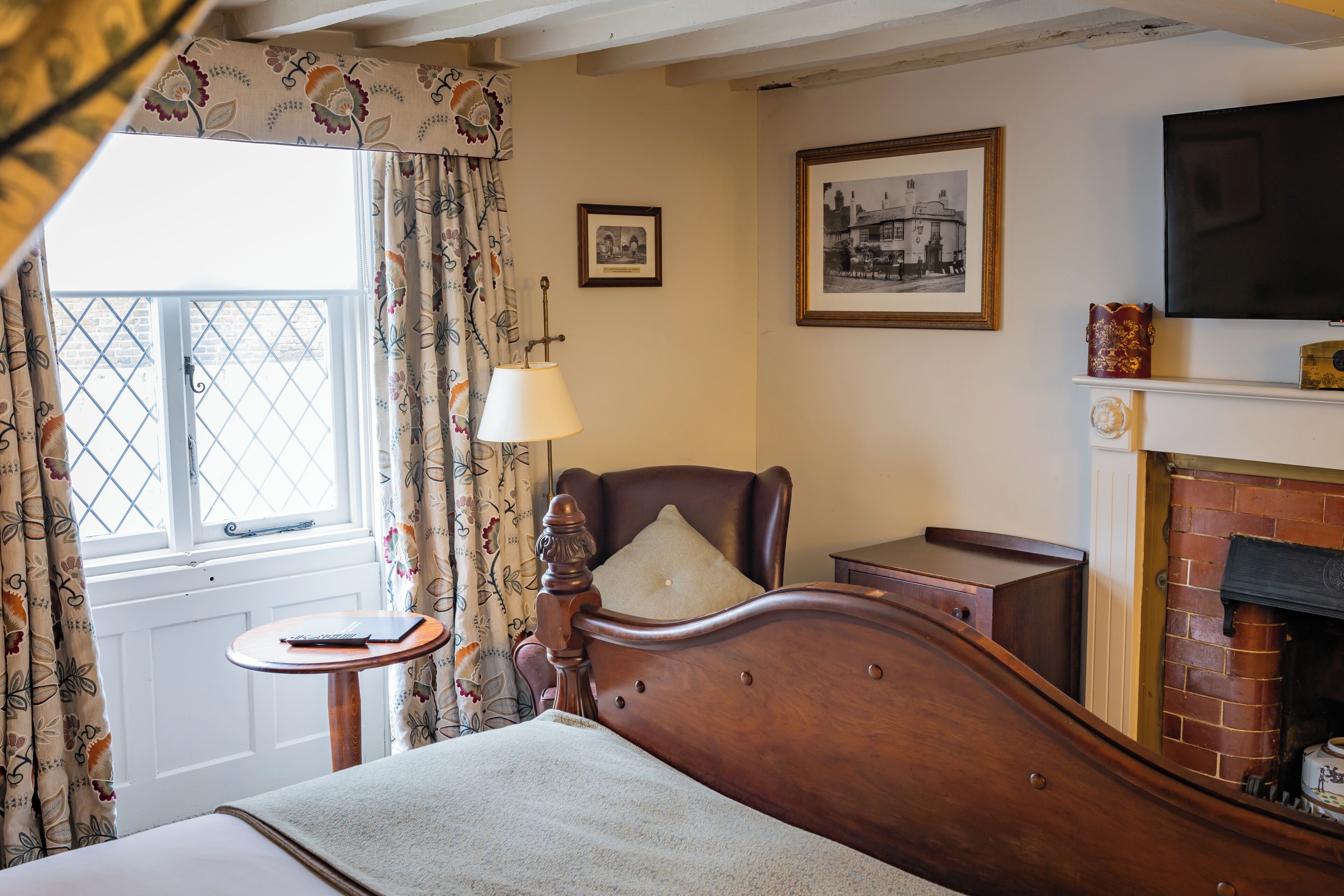 A bedroom with a wooden bed, a bedside table with a lamp, a fireplace, a window with curtains, and a framed picture on the wall.