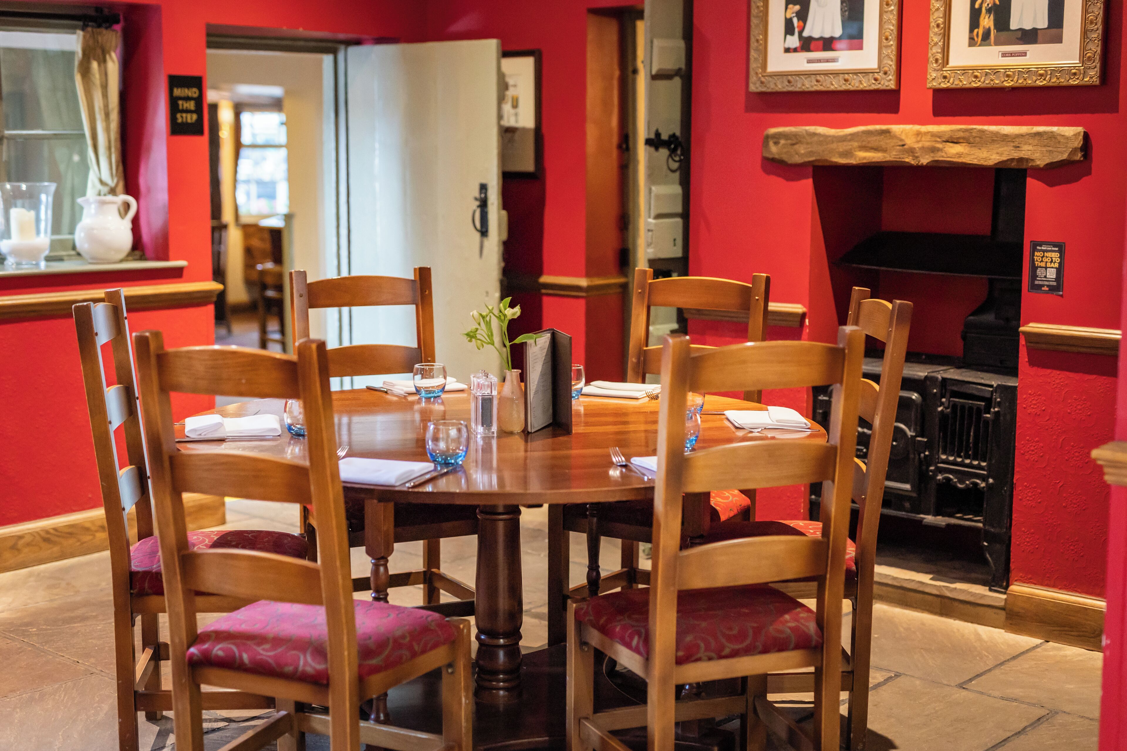 A dining area with a wooden table and chairs, a vase with flowers, and a red wall with framed pictures.