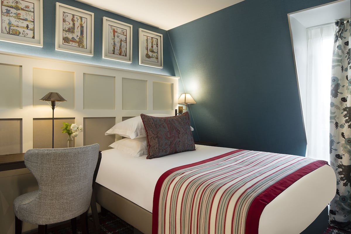 A hotel room with a bed, a chair, a desk, and framed artwork on the wall.