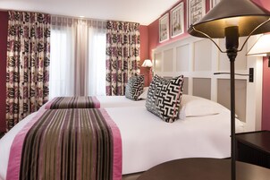 Camera d'albergo con un letto grande, due cuscini a motivo geometrico, una lampada da comodino e una finestra con tende leggere.